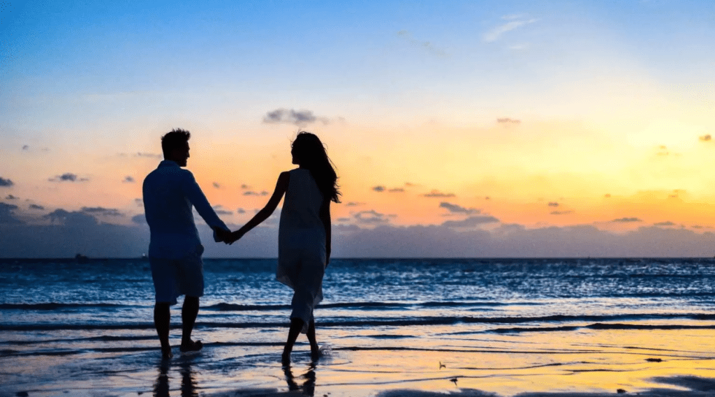 A couple holding hands on the beach at sunset, enjoying a romantic moment together amidst the beautiful scenery.