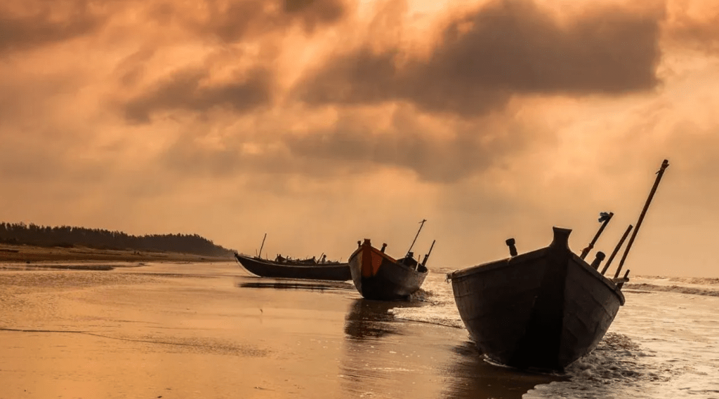 Three boats on a sandy beach of Odisha, illuminated by the warm hues of a sunset.
