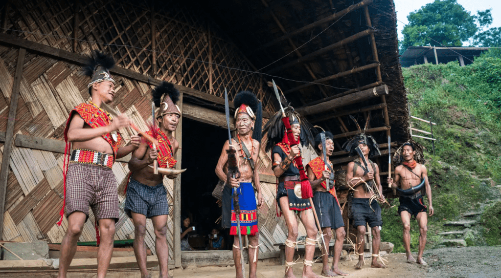 A diverse group of individuals wearing traditional attire pose in front of a rustic hut.