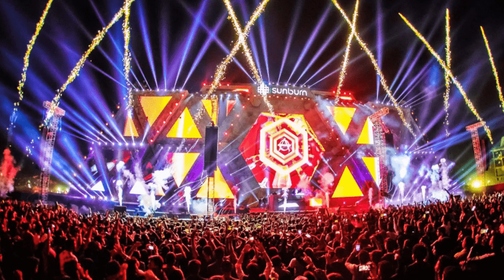 A vibrant music festival with a massive crowd, illuminated by dazzling fireworks in the background.