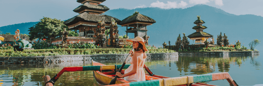 A woman paddles a boat on the water, with a temple visible in the background against a clear sky.