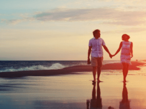 wo people walk hand in hand along a sandy beach, with gentle waves lapping at the shore in the background.