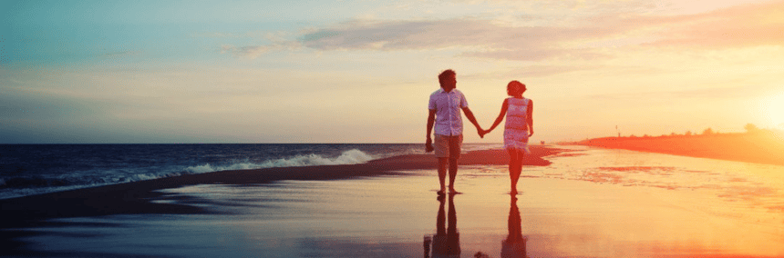 wo people walk hand in hand along a sandy beach, with gentle waves lapping at the shore in the background.