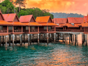 A row of wooden huts on stilts beside calm water, reflecting the serene landscape and clear blue sky.