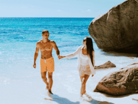 A couple walks hand in hand along the seychelle's beach, with gentle waves lapping at the shore in the background.