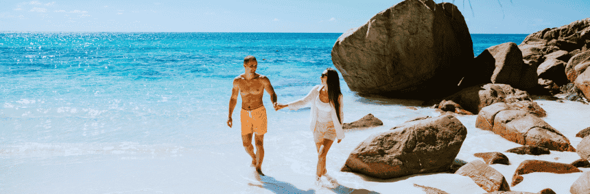 A couple walks hand in hand along the seychelle's beach, with gentle waves lapping at the shore in the background.