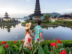 Couple holding hands at Ulun Danu Beratan Temple on Lake Bratan, Bali, with red flowers in front