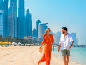 A couple strolls along the beach with a Dubai skyline visible in the background under a clear blue sky.