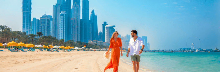 A couple strolls along the beach with a Dubai skyline visible in the background under a clear blue sky.